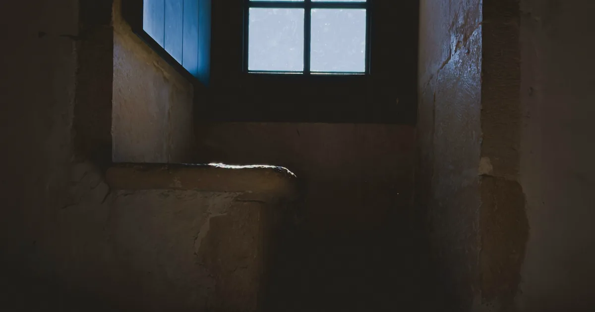 Étroite fenêtre au fond d’une pièce de pierre sombre, avec un rai de lumière sur le sol